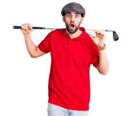 Young handsome man with beard playing golf holding club and ball scared and amazed with open mouth for surprise, disbelief face
