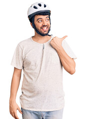 Young hispanic man wearing bike helmet smiling with happy face looking and pointing to the side with thumb up.