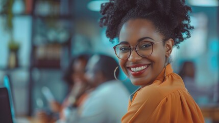 Fototapeta premium The image features a young, smiling African-American woman with glasses and an orange top. She is in a modern office setting, likely working on her laptop.