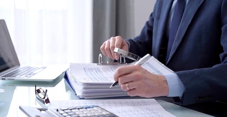 Businessman using a calculator and magnifying glass while analyzing financial documents at desk in modern office. Audit and taxes in business