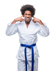 Afro american man wearing karate kimono over isolated background covering ears with fingers with annoyed expression for the noise of loud music. Deaf concept.