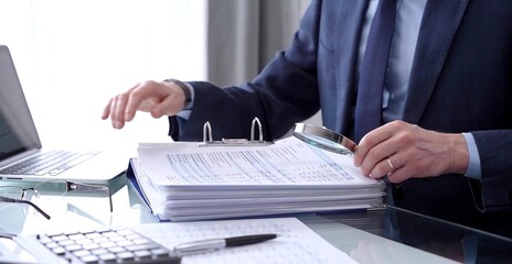 Businessman or auditor is looking through binders with papers while using a calculator and magnifying glass for financial documents analyzing. Audit and taxes concept