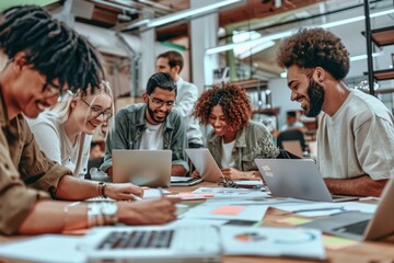 Smiling diverse team working together in a creative office environment, using laptops and papers to develop advertising ideas. High-resolution image showcasing teamwork and innovation.