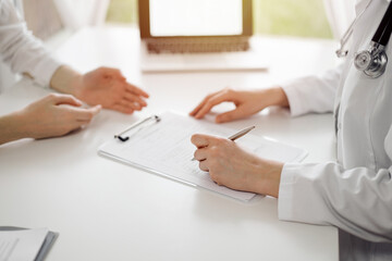 Doctor and patient sitting near each other at the white desk in clinic. Female physician is listening filling up a records form. Medicine concept