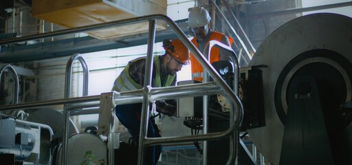 Two professional heavy industry engineers in protective uniforms and helmets at manufacturing facility or factory. Male worker talks and shows machinery equipment to mature supervisor. Slow motion.