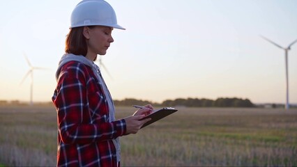 Adult woman engineer wearing white cask is taking notes on a clipboard on a field with wind turbines