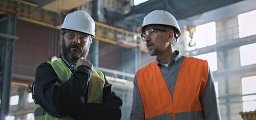 Two professional heavy industry engineers in safety uniform and hard hats discuss work process at manufacturing factory. Mature supervisor talks to adult worker at plant or workshop. Slow motion.