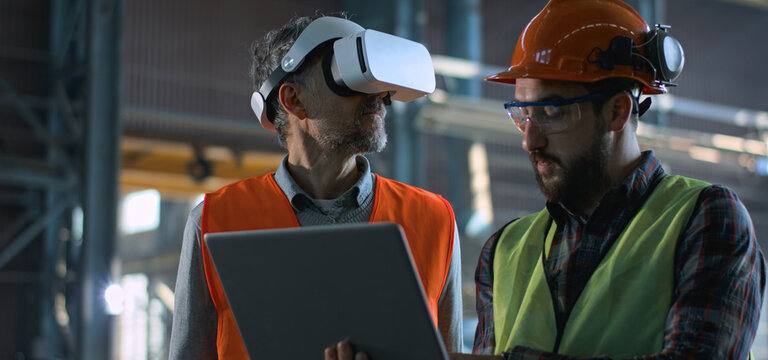 Two professional industry technicians wearing safety uniform and hardhats stand at manufacturing factory, use laptop and AR headset. Mature engineer and adult employee work in warehouse. Slow motion.