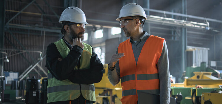 Two professional heavy industry engineers in safety uniform and hard hats discuss work process at manufacturing factory. Mature supervisor talks to adult worker at plant or workshop. Slow motion.