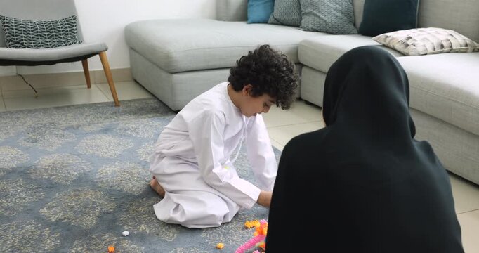 Cute Arab son boy in white ethnic cloth and mother in black abaya playing with toy construction bricks on home floor, stacking colorful blocks, having fun, enjoying family game, entertainment
