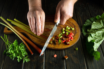 A chef uses his hands and a knife to cut rhubarb stalks on a cutting board. Preparing a vegetable mix on a breakfast table.