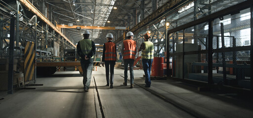 Back view of four heavy industry technicians in uniform and helmets discussing production process, walking at manufacturing plant. Male and female inspectors talk to employees in factory. Slow motion.