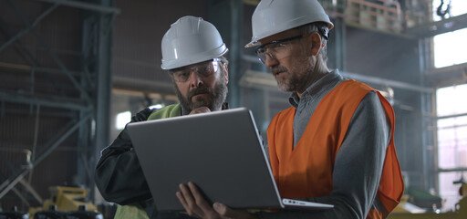Two professional heavy industry workers, engineers wearing protective uniform and hardhats discuss work process using laptop computer while standing in manufacturing factory or warehouse. Slow motion.
