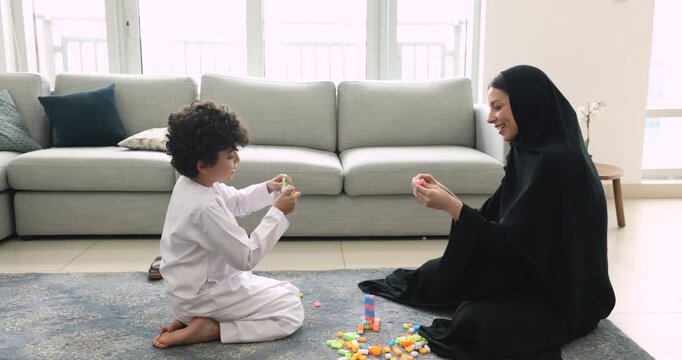 Happy Arabic mom in black Muslim clothes playing with son kid in white thobe at home, sitting on floor, constructing tower from building blocks, talking to child, smiling, enjoying family playtime