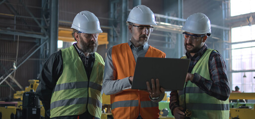 Three professional heavy industry technicians in safety uniform and helmets talk about production. Mature inspector uses laptop standing with colleagues in manufacturing plant or factory. Slow motion.