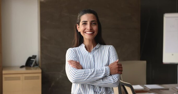 Serious beautiful young Hispanic business leader woman standing in office, folding arms in confidence gesture, getting happy, looking at camera with toothy smile. CEO, executive, director head shot