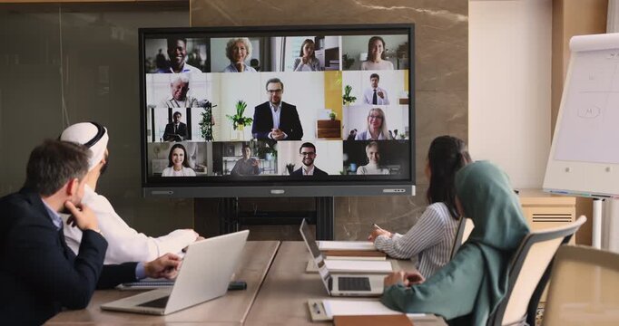 Diverse team of Arabic end European businesspeople talking to colleagues on online conference call, looking at large display, electronic board split screen, meeting on Internet for group communication