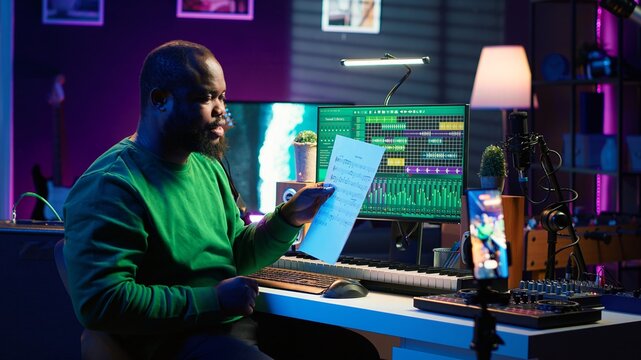 Artist pointing at musical sheet notation to explain notes on piano, teaching people to play synthesizer in home studio. Composer musician playing midi controller during online master class. Camera A.