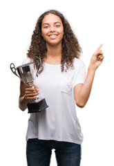 Young hispanic woman holding trophy very happy pointing with hand and finger to the side