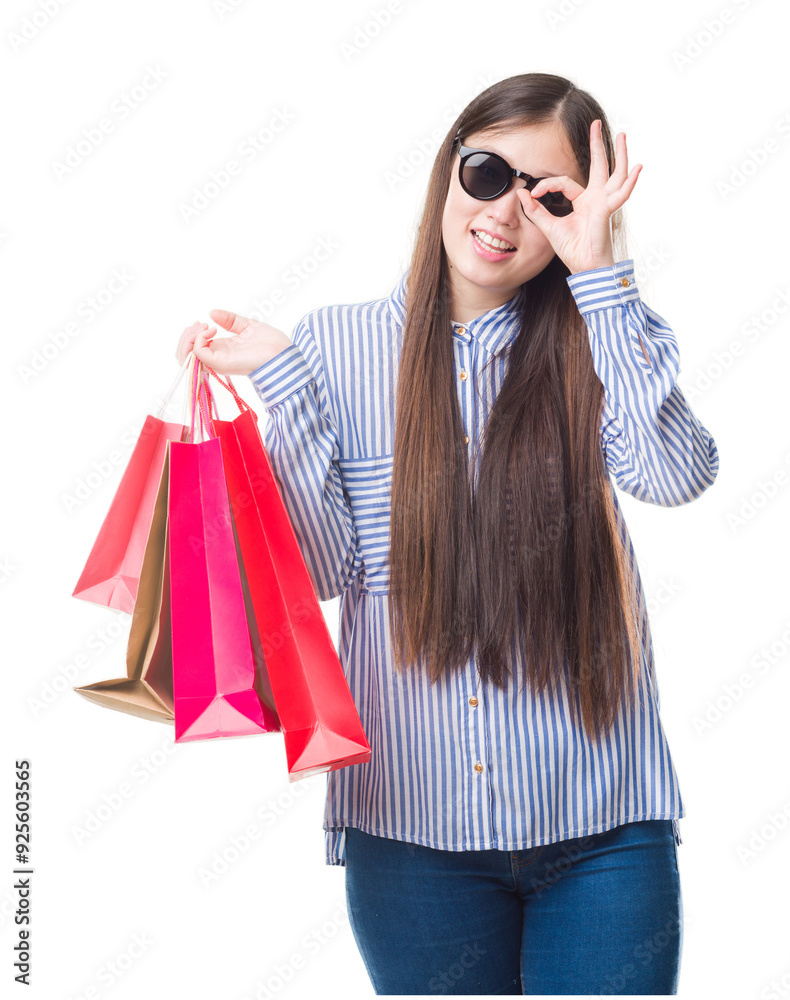 Wall mural Young Chinese woman over isolated background holding shopping bags on sales with happy face smiling doing ok sign with hand on eye looking through fingers