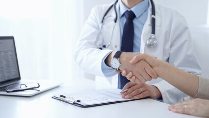 Doctor and a patient are shaking hands. The physician is wearing a white medical coat over a blue shirt. Medical service