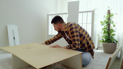 Young man struggles to correctly assemble a white wooden cabinet for the baby with the help of a screwdriver. Future father prepares the room of the newborn. Cozy picture from a bright room