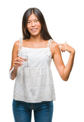 Young asian woman drinking glass of water over isolated background with surprise face pointing finger to himself