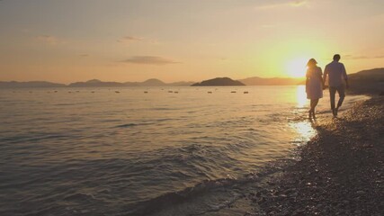 A couple walks hand in hand along the beach at sunrise, with the golden light reflecting on the calm sea and distant islands, creating a serene and romantic atmosphere.