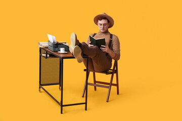 Young man reading book with cup of coffee and vintage typewriter on table against yellow background