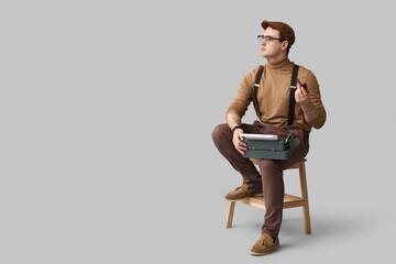 Young man with vintage typewriter smoking pipe on chair against grey background