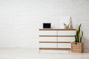 Wooden chest of drawers with laptop, houseplants and decor near white brick wall