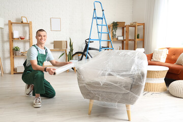 Male worker wrapping armchair with stretch film in room