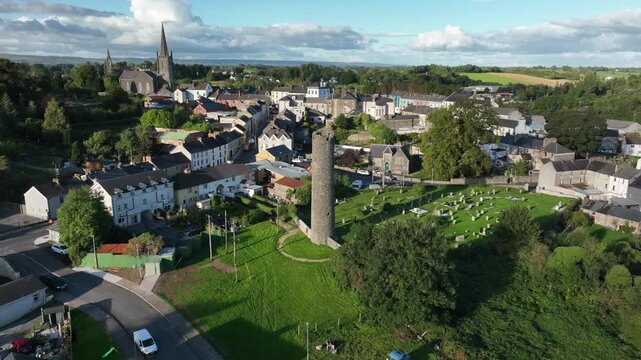 Clones Town, County Monaghan, Ireland, April 2023. Drone pulls backward ascending from the historic Round Tower casting long shadows over cemetery in the country town.