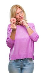 Middle age blonde woman eating pink donut over isolated background serious face thinking about question, very confused idea