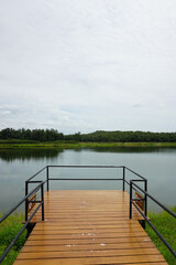 A tranquil scene of a wooden pier extending into a calm lake. The pier has a metal railing and offers a peaceful view of the surrounding natural scenery. Lush green hills and a cloudy sky complete the