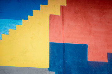 A colorful wall with a yellow stair step and blue and red squares. The wall is painted in a way that it looks like a staircase