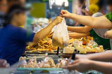 A woman is handing a plastic bag to another woman. The bag is filled with food. The scene is set in a market where people are buying and selling food