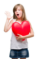 Young blonde toddler holding a red heart very happy and excited, winner expression celebrating victory screaming with big smile and raised hands