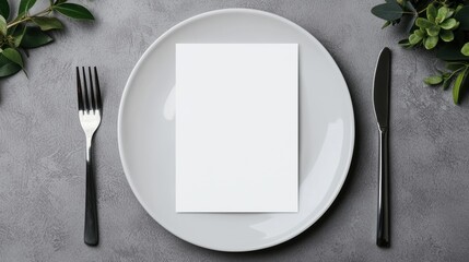 Minimalist Table Setting with Blank Card on White Plate, Fork, and Knife on Gray Background with Greenery