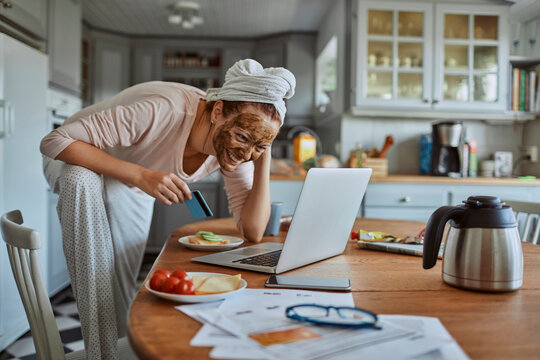 Woman with face mask enjoying breakfast and coffee while working on laptop in kitchen