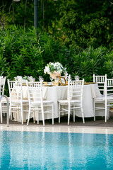 Outdoor Elegant Event by the Pool: A beautifully set round dining table with white chairs and a floral centerpiece, situated next to a swimming pool, perfect for a garden party, wedding