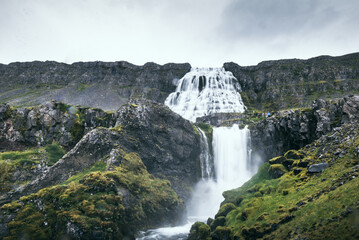 Majestic Dynjandi waterfall cascades over rocky cliffs in Iceland’s Westfjords, surrounded by rugged landscapes and lush green moss. Overcast sky enhances the dramatic mood.