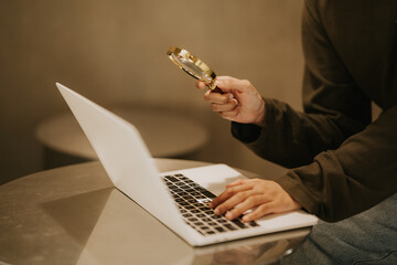 Digital Scrutiny:  A person meticulously analyzes data on a laptop, magnifying glass in hand, in a...