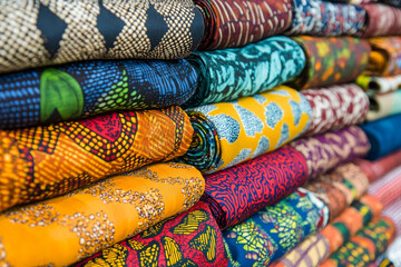 A close-up of vibrant, patterned fabrics stacked neatly in a market, showcasing traditional textile designs and diverse color palettes.
