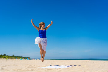 Obraz premium Young woman in blue swimsuit, white pareo and sunglasses jumping high up on white sand on beautiful beach against blue sky on sunny day. Energy and power on beach.