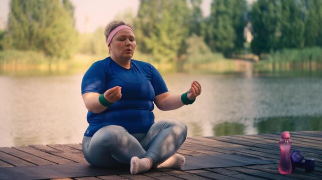 Overweight woman meditating in lotus asana pose by a lake, outdoor fitness 