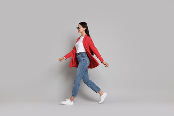 Stylish woman in red jacket, jeans and sunglasses walking on gray background