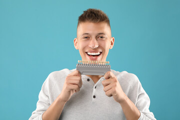 Happy young man with teeth color samples on light blue background. Dental veneers