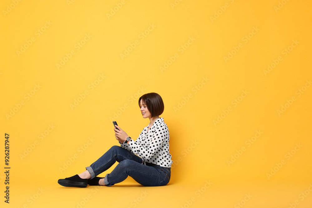 Wall mural Smiling woman with smartphone sitting on yellow background. Space for text