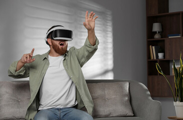 Emotional man using virtual reality headset on sofa at home. Space for text